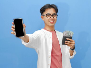 Elinde para dolu bir cüzdanla gülümseyen Asyalı genç bir adamın portresi ve mavi arka planda izole edilmiş boş bir cep telefonu ekranı..