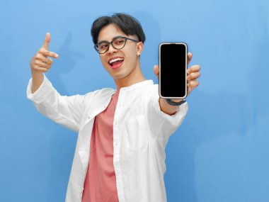 Sıradan giyinmiş genç bir Asyalı 'nın portresi mavi arka planda izole edilmiş boş bir cep telefonu ekranını işaret ediyor..