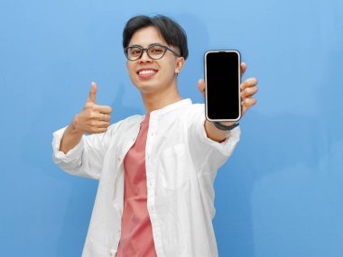 Heyecanlı Asyalı adam, günlük giysiler içinde boş telefon ekranı gösteriyor, mavi arka planda mutlu bir ifadeyle başparmak kaldırıyor..