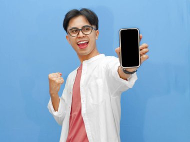 Sıradan kıyafetler içinde genç bir Asyalı adamın portresi boş bir cep telefonu ekranı tutuyor, başarıyı mavi arka planda heyecanlı bir ifadeyle kutluyor..