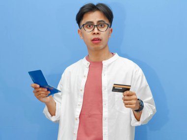 Tasarruf defteri ve kredi kartı tutan kafası karışmış Asyalı bir adam mavi arka planda şok olmuş bir ifadeyle mali içerik ve reklamlar için mükemmel..
