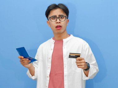 Tasarruf defteri ve kredi kartı tutan kafası karışmış Asyalı bir adam mavi arka planda şok olmuş bir ifadeyle mali içerik ve reklamlar için mükemmel..