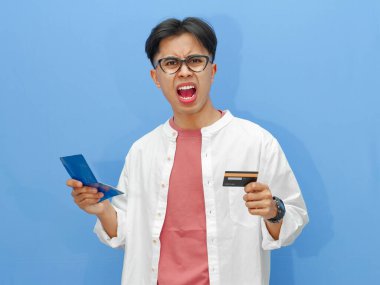 Tasarruf defteri ve kredi kartı tutan kafası karışmış Asyalı bir adam mavi arka planda şok olmuş bir ifadeyle mali içerik ve reklamlar için mükemmel..