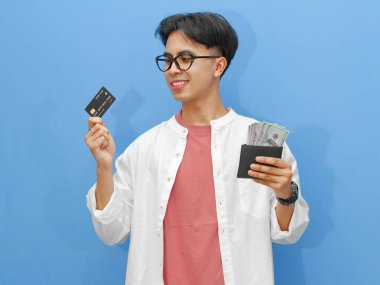 Elinde para dolu bir cüzdan ve gülümseyen mutlu bir ifadeyle banka kartı tutan genç bir Asyalı adam, mavi bir arka planda izole edilmiş, reklam için mükemmel..
