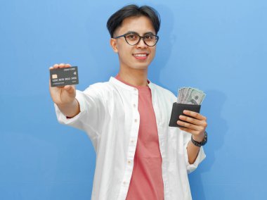 Elinde para dolu bir cüzdan ve gülümseyen mutlu bir ifadeyle banka kartı tutan genç bir Asyalı adam, mavi bir arka planda izole edilmiş, reklam için mükemmel..