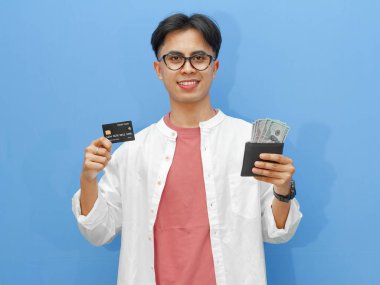 Elinde para dolu bir cüzdan ve gülümseyen mutlu bir ifadeyle banka kartı tutan genç bir Asyalı adam, mavi bir arka planda izole edilmiş, reklam için mükemmel..