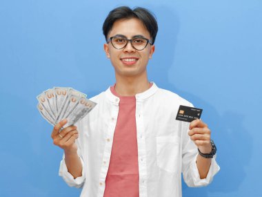 Asyalı genç bir adam elinde banka kartı ve mutlu bir ifadeyle mavi bir arka planda izole edilmiş nakit para tutuyor. Finansal, bankacılık ya da iş promosyonları için mükemmel.