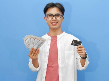 Asyalı genç bir adam elinde banka kartı ve mutlu bir ifadeyle mavi bir arka planda izole edilmiş nakit para tutuyor. Finansal, bankacılık ya da iş promosyonları için mükemmel.