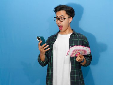 Asyalı genç adam para ve telefon taşıyan sıradan bir tişört giymiş, komik bir surat ifadesi sergiliyor..