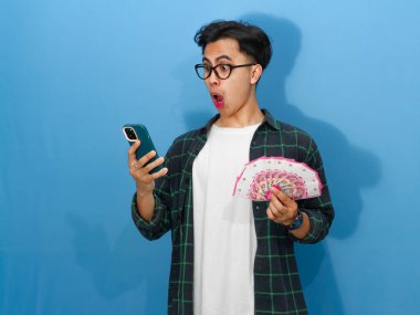 Asyalı genç adam para ve telefon taşıyan sıradan bir tişört giymiş, komik bir surat ifadesi sergiliyor..