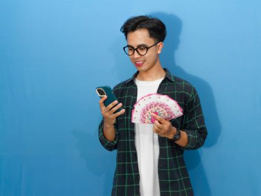 Sıradan gömlekli genç bir Asyalı elinde banknotlar ve gülümseyen bir cep telefonu kullanıyor..