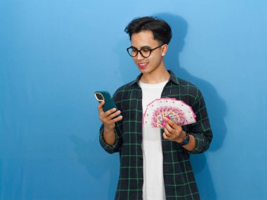 Sıradan gömlekli genç bir Asyalı elinde banknotlar ve gülümseyen bir cep telefonu kullanıyor..