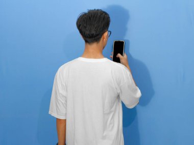 Mavi arka planda cep telefonu kullanan beyaz tişörtlü genç bir Asyalı. Reklam ve pazarlama için ideal..