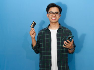 Banka kartı ve cep telefonu tutarak poz veren sıradan bir tişört giyen Asyalı genç bir adamın portresi kendine güvenen yüz ifadesini gösteriyor..