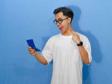 Beyaz tişörtlü genç Asyalı bir adamın portresi banka tasarruf defterini tutarken heyecanlı bir ifadeyle başarıyı kutluyor..