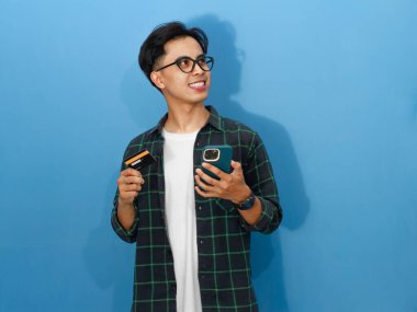 Heyecanlı Asyalı genç adam elinde bir kredi kartı ve cep telefonu tutarken gülümsüyor, yukarı bakıyor, finans ve yaşam tarzı temaları için mükemmel..