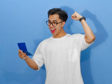 Beyaz tişörtlü genç Asyalı bir adamın portresi banka tasarruf defterini tutarken heyecanlı bir ifadeyle başarıyı kutluyor..