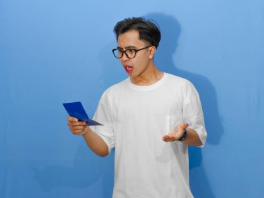 Beyaz tişörtlü kafası karışmış Asyalı genç bir adamın portresi, elinde bir banka tasarruf defteri tutarken şok edici bir ifade sergiliyor..