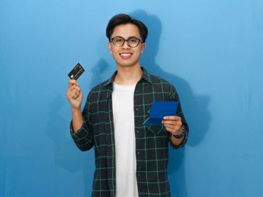 Sıradan tişörtlü, kendine güvenen genç Asyalı bir adam elinde kredi kartı ve tasarruf defteri tutarken gülümsüyor ve kameraya bakıyor..