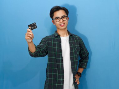 Sıradan gömlekli, kendine güvenen genç Asyalı bir adam elinde kredi kartı tutarken gülümsüyor ve olumlu bir ifade sergiliyor..
