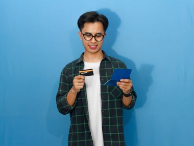 Genç Asyalı adam elinde kredi kartı tutarken mutlu bir şekilde tasarruf defterini kontrol ediyor ve olumlu bir mali tavır sergiliyor..