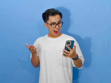 Beyaz tişörtlü genç bir Asyalı adamın portresi. Elinde cep telefonu tutarken şaşırmış bir ifade var. Reklam veya promosyon için mükemmel..