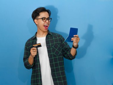 Asyalı heyecanlı genç adam başarıyı kredi kartı ve banka tasarruf kitabıyla kutluyor, neşe ve başarı sergiliyor..