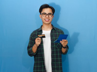 Sıradan tişörtlü, kendine güvenen genç Asyalı bir adam elinde kredi kartı ve tasarruf defteri tutarken gülümsüyor ve kameraya bakıyor..