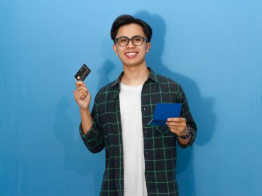 Sıradan tişörtlü, kendine güvenen genç Asyalı bir adam elinde kredi kartı ve tasarruf defteri tutarken gülümsüyor ve kameraya bakıyor..