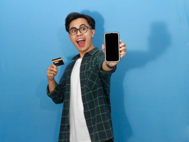 Sıradan gömlekli, Asyalı genç bir adam boş bir cep telefonu ekranı gösteriyor ve elinde internet üzerinden yapılan ödemelerin altını çizen bir banka kartı var..