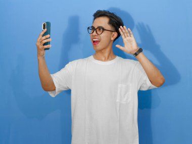 Beyaz tişörtlü, cep telefonuyla poz veren heyecanlı Asyalı genç bir adam video konuşması yapıyor. İletişim ya da teknoloji temaları için mükemmel.