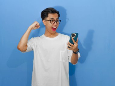 Beyaz tişörtlü heyecanlı Asyalı genç adam bir cep telefonuna bakarken başarıyı kutluyor, neşe ve başarıyı ifade ediyor..