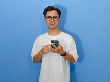 Elinde cep telefonu tutan beyaz tişörtlü gülümseyen Asyalı genç bir adamın portresi. Reklam, iletişim ve teknoloji kavramları için mükemmel..