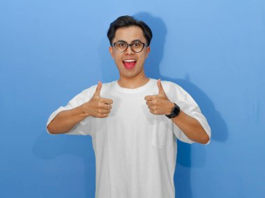 Beyaz tişörtlü heyecanlı genç Asyalı adam, kameraya gülümsüyor ve başparmak kaldırıyor, olumlu mesajlar için mükemmel..