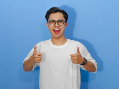 Beyaz tişörtlü heyecanlı genç Asyalı adam, kameraya gülümsüyor ve başparmak kaldırıyor, olumlu mesajlar için mükemmel..