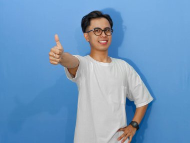 Beyaz tişörtlü heyecanlı genç Asyalı adam, kameraya gülümsüyor ve başparmak kaldırıyor, olumlu mesajlar için mükemmel..