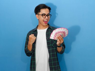 Sıradan gömlekli genç bir Asyalı 'nın portresi para tutuyor, yumruklarını sıkıyor ve başarıyı heyecanlı bir ifadeyle kutluyor..