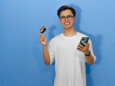 Beyaz tişörtlü, elinde banka kartı ve cep telefonu olan heyecanlı Asyalı genç adam, mutlu bir şekilde gülümsüyor, ödeme, alışveriş ve teknoloji kavramları için mükemmel..