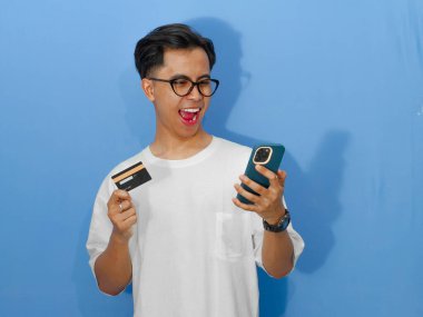 Beyaz tişörtlü heyecanlı Asyalı genç adam elinde bir banka kartı ve cep telefonu tutarken komik bir şok ifadesi gösteriyor..