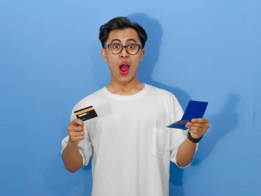 Beyaz tişörtlü genç bir Asyalı elinde banka kartı ve tasarruf defteri tutarken komik ve şaşırmış bir ifade sergiliyor..