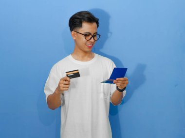 Beyaz tişörtlü gülümseyen Asyalı bir adamın portresi, elinde banka kartı ve tasarruf defteri var, finansal başarıyı temsil ediyor..