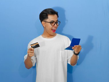 Beyaz tişörtlü genç bir Asyalı elinde banka kartı ve tasarruf defteri tutarken komik ve şaşırmış bir ifade sergiliyor..