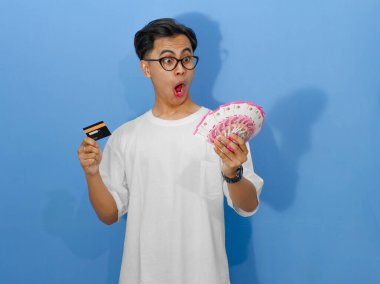 Beyaz tişörtlü heyecanlı Asyalı genç adam elinde bir banka kartı ve para tutarken komik bir sürpriz ifade sergiliyor..