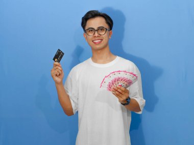 Beyaz tişörtlü genç bir Asyalı 'nın portresi. Banka kredi kartı ve nakit para taşırken mutlu bir şekilde gülümsüyor..