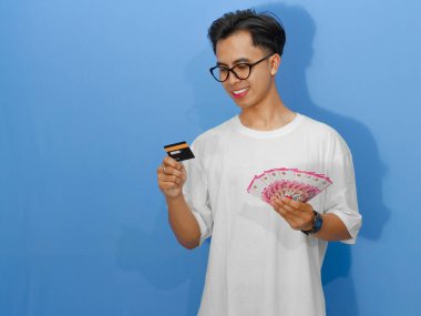 Beyaz tişörtlü genç bir Asyalı 'nın portresi. Banka kredi kartı ve nakit para taşırken mutlu bir şekilde gülümsüyor..