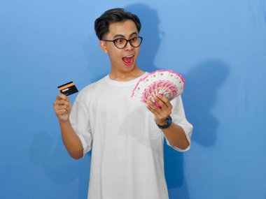 Beyaz tişörtlü heyecanlı Asyalı genç adam elinde bir banka kartı ve para tutarken komik bir sürpriz ifade sergiliyor..