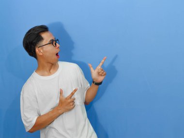 Beyaz tişörtlü heyecanlı genç Asyalı adam yan tarafı gösterirken komik bir sürpriz ifade gösteriyor..