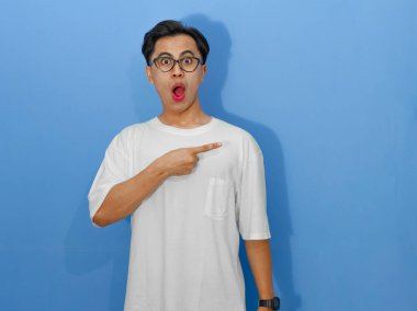 Beyaz tişörtlü heyecanlı genç Asyalı adam yan tarafı gösterirken komik bir sürpriz ifade gösteriyor..