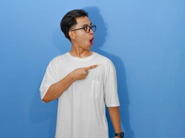 Beyaz tişörtlü heyecanlı genç Asyalı adam yan tarafı gösterirken komik bir sürpriz ifade gösteriyor..