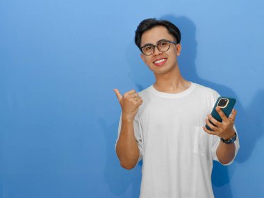 Beyaz tişörtlü, cep telefonuyla poz veren ve baş parmağıyla işaret eden mutlu bir Asyalı genç adam portresi..